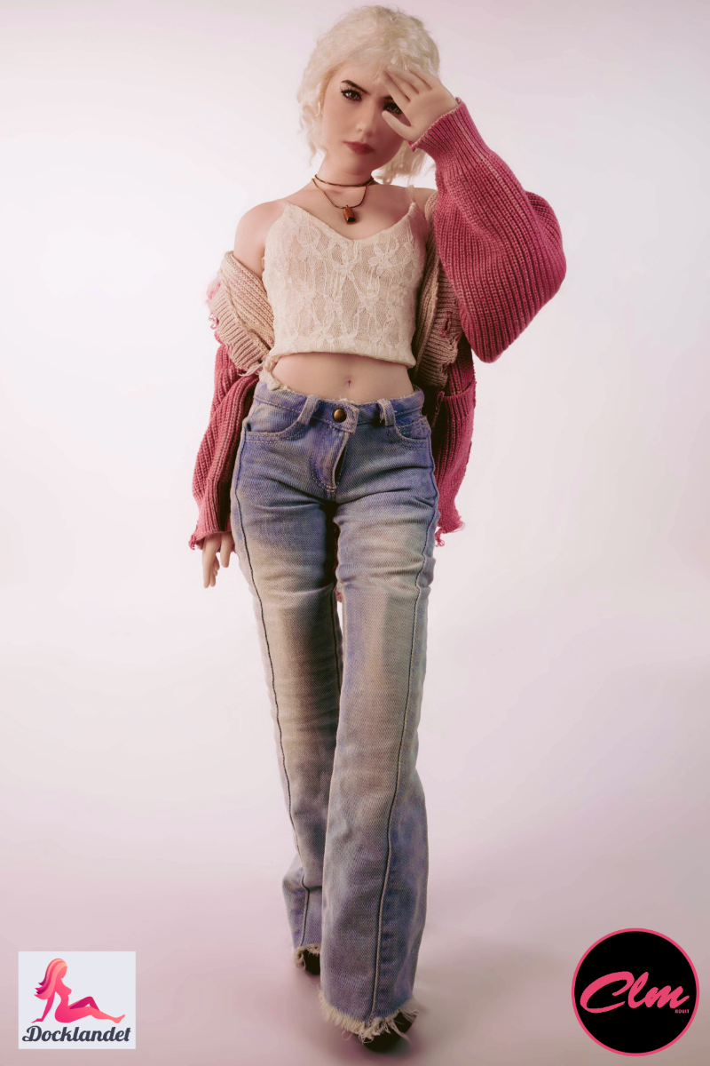 sexdukke Tara realistisk mini 60 cm in silikone, blondine dukke i jeans, crop top og pink jakke, stående oprejst mod lys studiebaggrund.